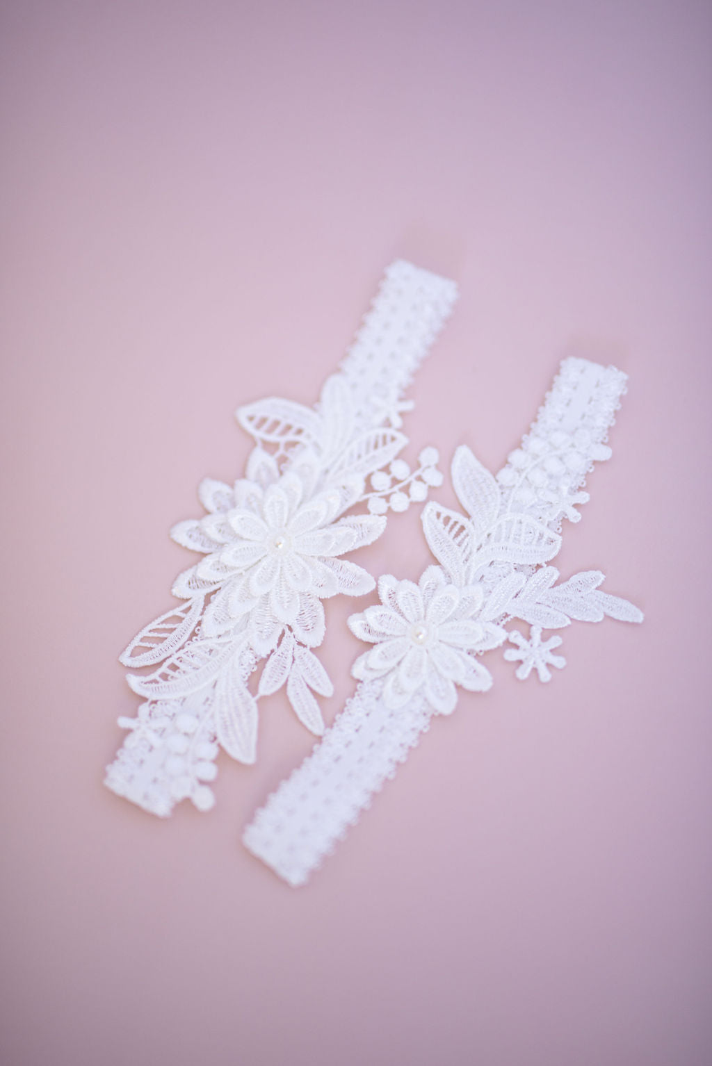 A pair of white lace garters with floral detailing and pearl accents on a pink background.
