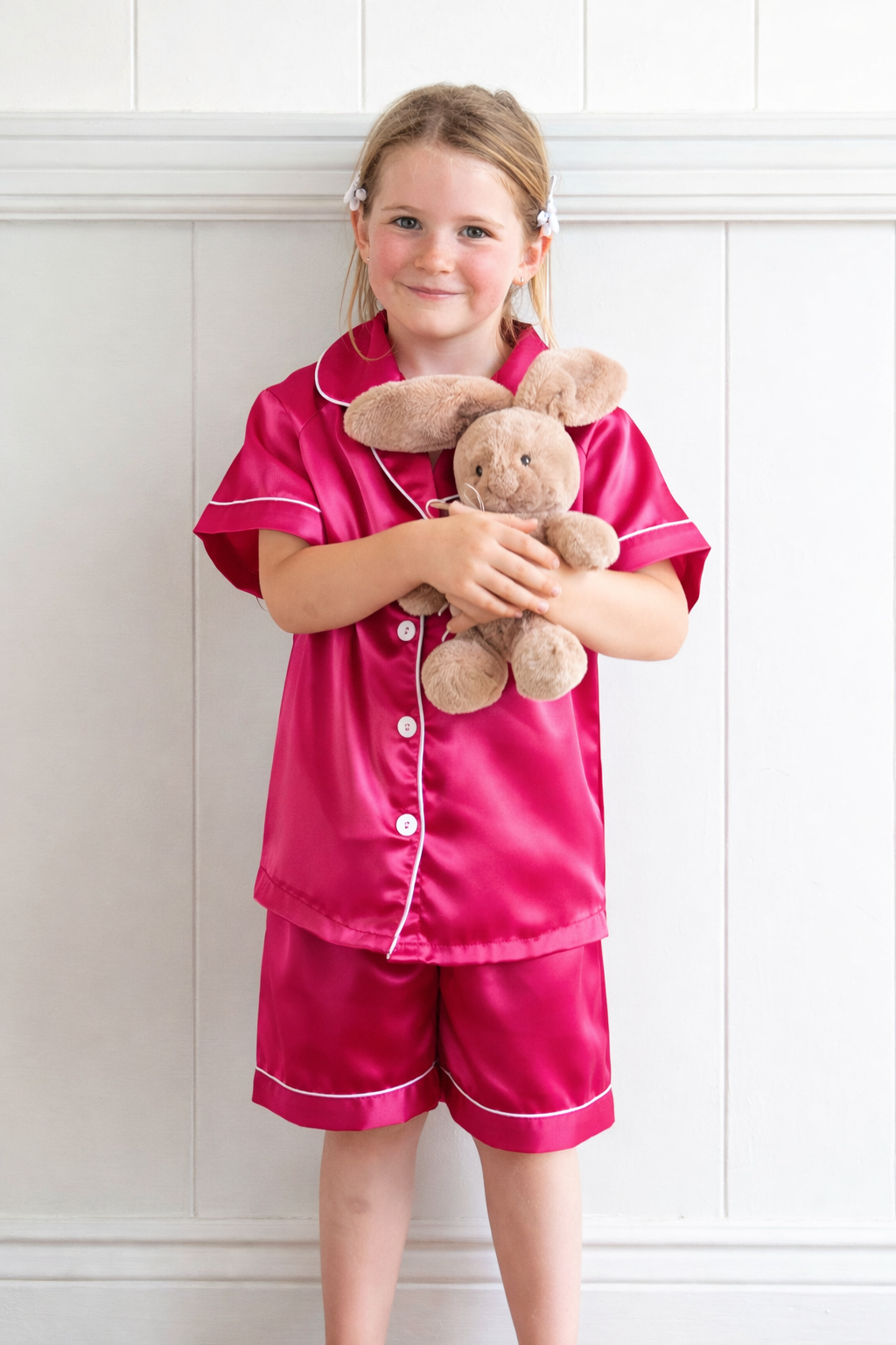 Young girl wearing bright red satin kids pyjamas with white piping, standing barefoot indoors and holding a soft teddy bunny
