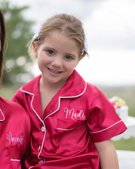 Smiling young girl wearing personalised bright red satin kids pyjamas with white piping and name embroidery, outdoor portrait
