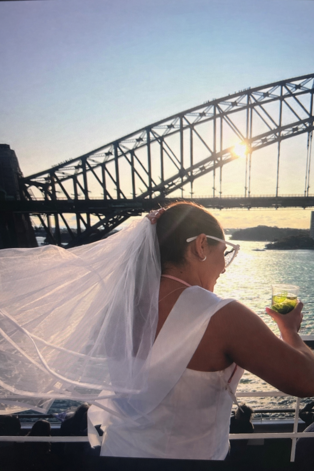 Person with a white garment, possibly a veil, standing by a waterfront with a bridge in the background.