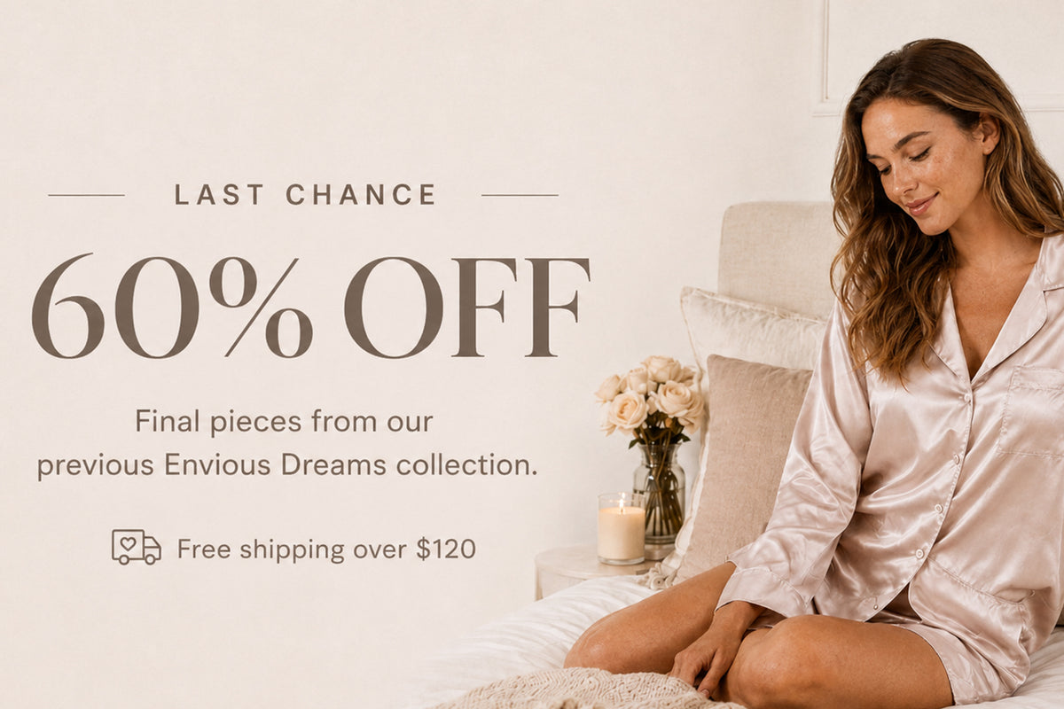 Woman wearing blush satin pyjamas seated on a neutral bed for the Envious Dreams final clearance sale.