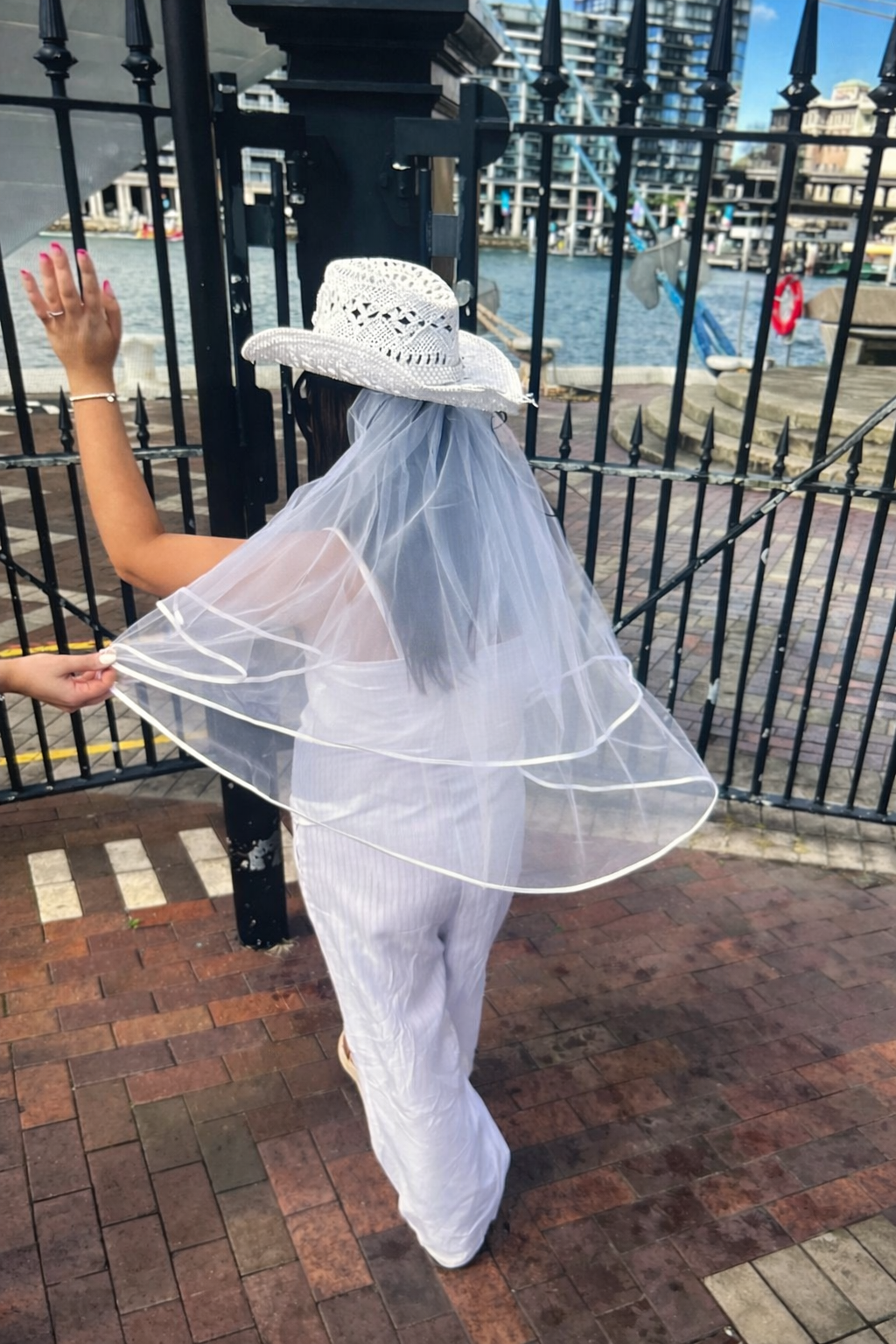 Back view of bride wearing a two-tier white bridal veil with satin edging and a white woven hat, standing by a waterfront city promenade.
